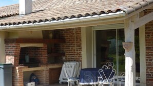 A patio with a brick fireplace, outdoor seating, and a covered area with a tiled roof. - Gite on closed wooded grounds (Castelferrus)