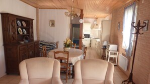A dining area with two upholstered chairs, a wooden cabinet with decorative plates, a dining table with a floral centerpiece, and a kitchen in the background. - Gite on closed wooded grounds (Castelferrus)