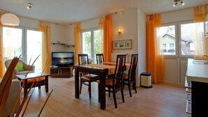 Een moderne woonkamer met een eetgedeelte, grote ramen met gordijnen, een televisie en een keuken.