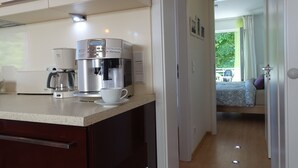 Een moderne keuken met een koffieapparaat en een kopje op het aanrecht. Een hal leidt naar een slaapkamer met een bed en een raam.