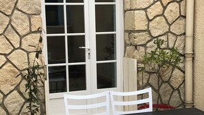 Un muro de piedra, una ventana con contraventanas blancas, una mesa con sillas y plantas en macetas.