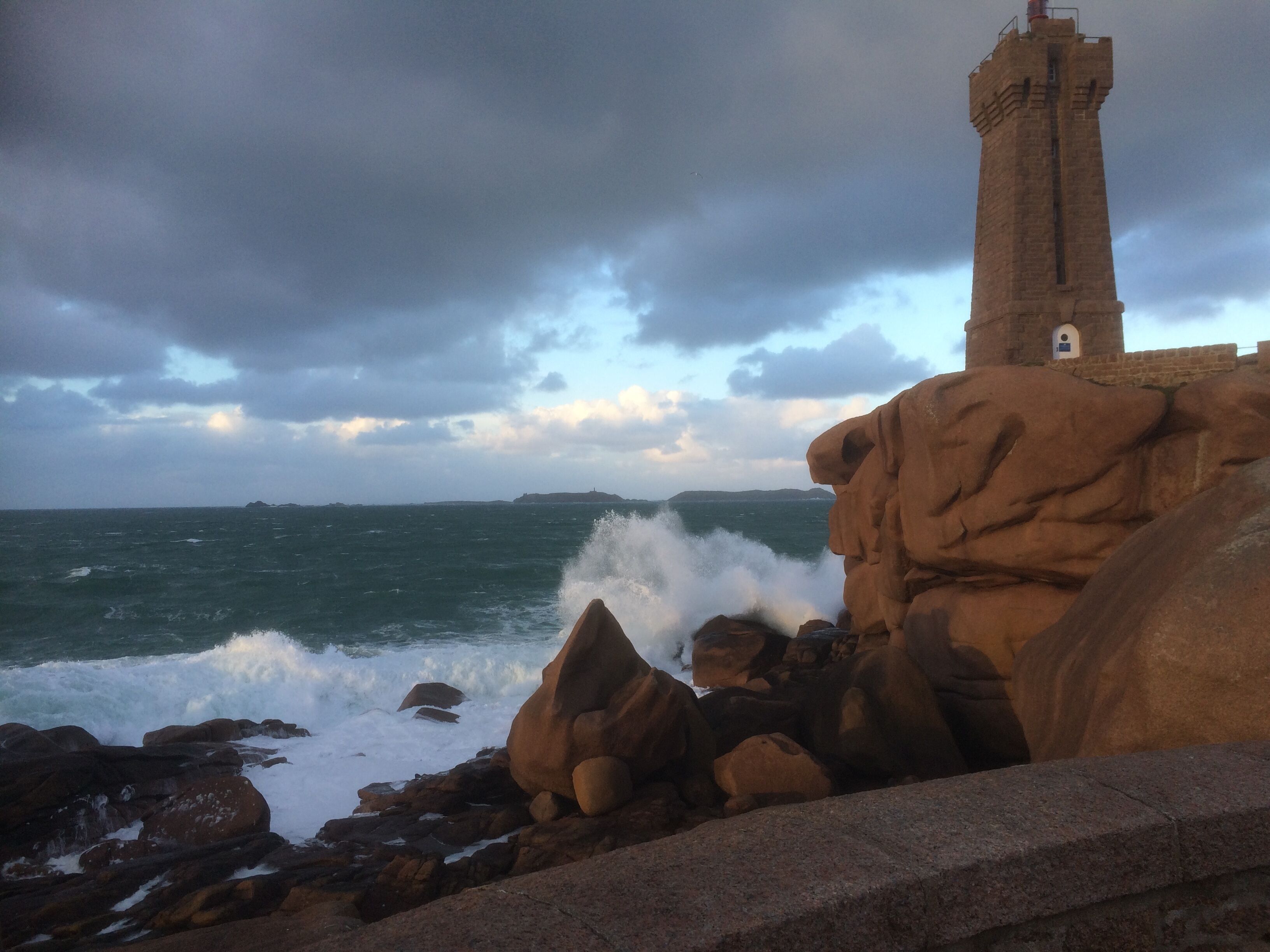 Paisaje costero con un faro sobre un promontorio rocoso, olas rompiendo contra las rocas y un cielo nublado.