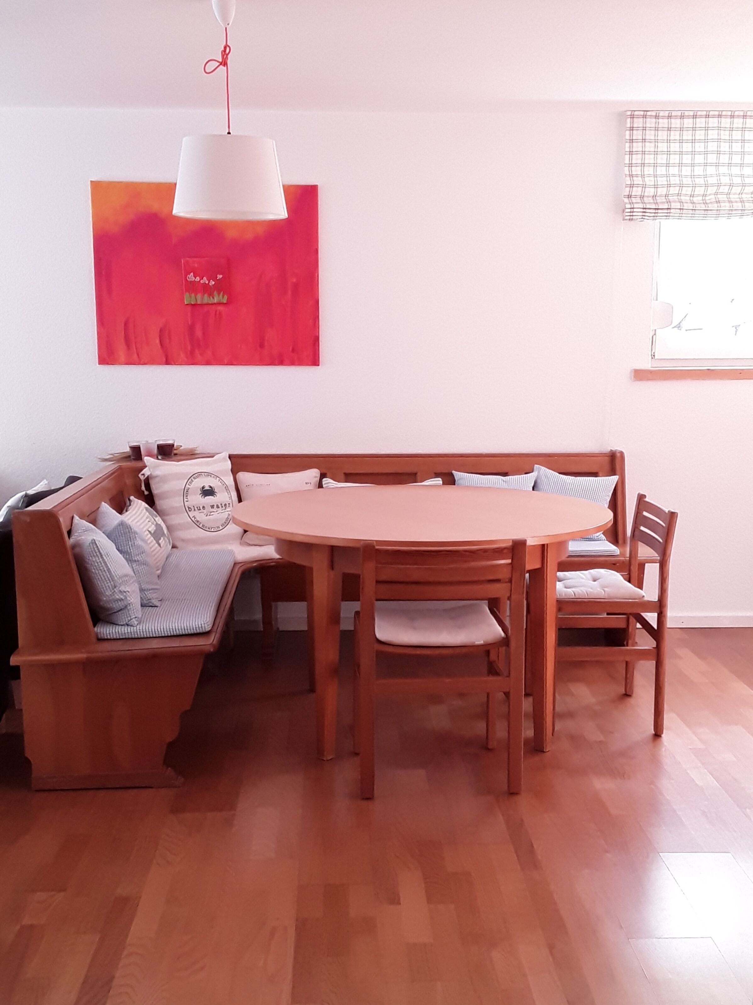 A dining area with a wooden table and benches, a hanging pendant light, and a red abstract painting on the wall.