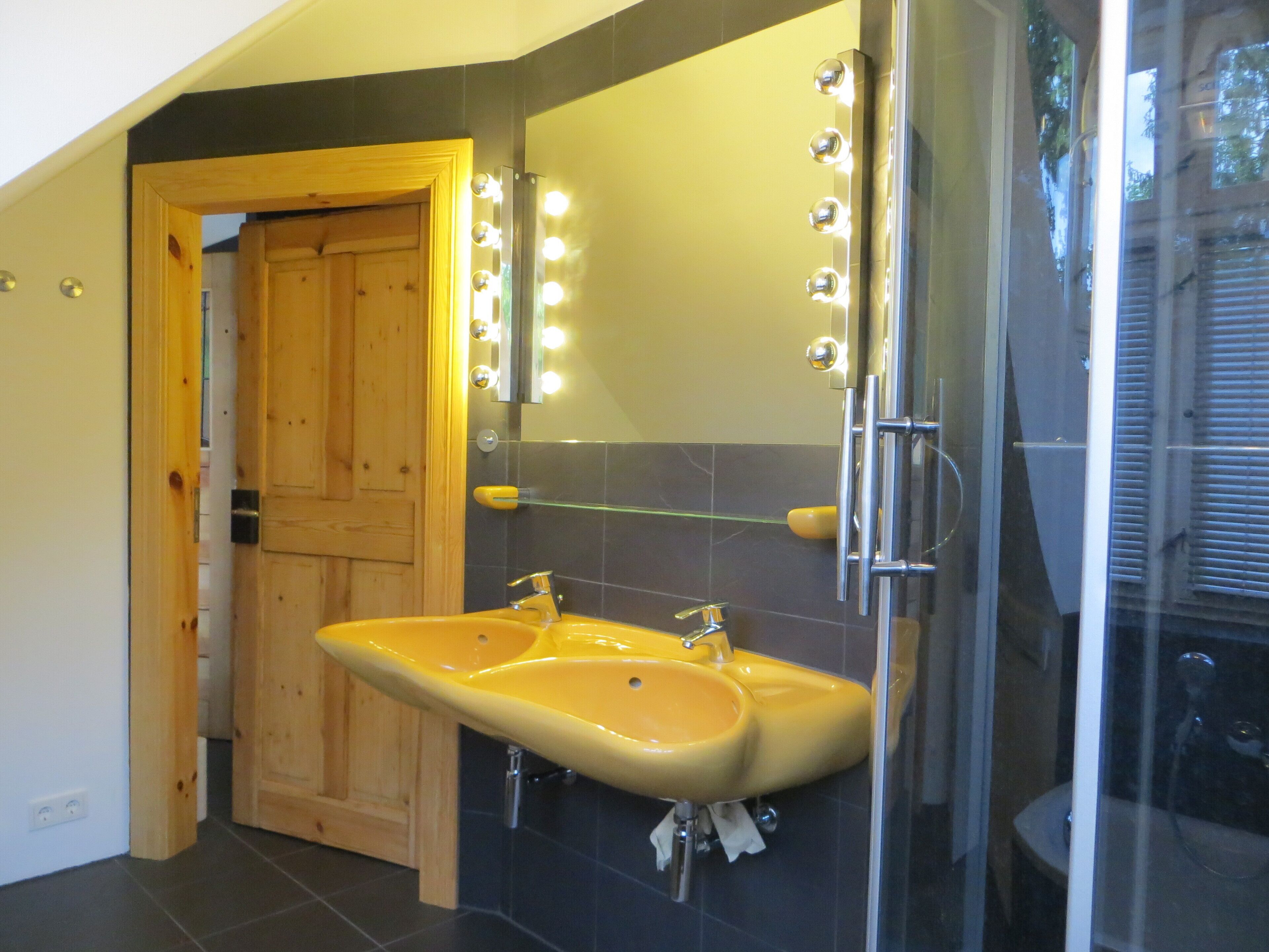 A bathroom with a yellow sink, a mirror with lights, a glass shower enclosure, and a wooden door.