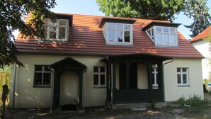 Une maison à deux étages, avec un toit recouvert de tuiles rouges, un porche à l’avant doté d’une balustrade noire et une petite porte d’entrée.