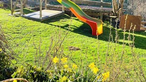 Una casita de juegos de madera con un tobogán en un área verde con árboles y colinas al fondo.