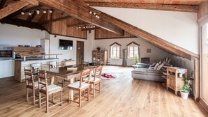 Un loft espacioso con techo alto, vigas de madera, zona de comedor con mesa larga y sillas, cocina y un rincón de estar acogedor con sofá y cojines.