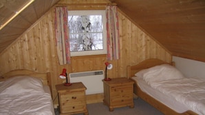 A small, cozy attic bedroom with two wooden beds, a window with curtains, and a radiator.