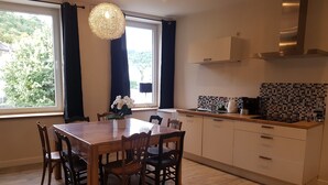 A modern kitchen with a wooden dining table and chairs, a large window with curtains, and a hanging light fixture. - Gîte Histoire d'Ardenne with sauna in the historic center of La Roche (La Roche-en-Ardenne)