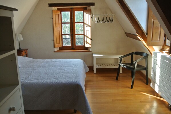 Un dormitorio con cama, una silla, una ventana con contraventanas de madera y un banco blanco.
