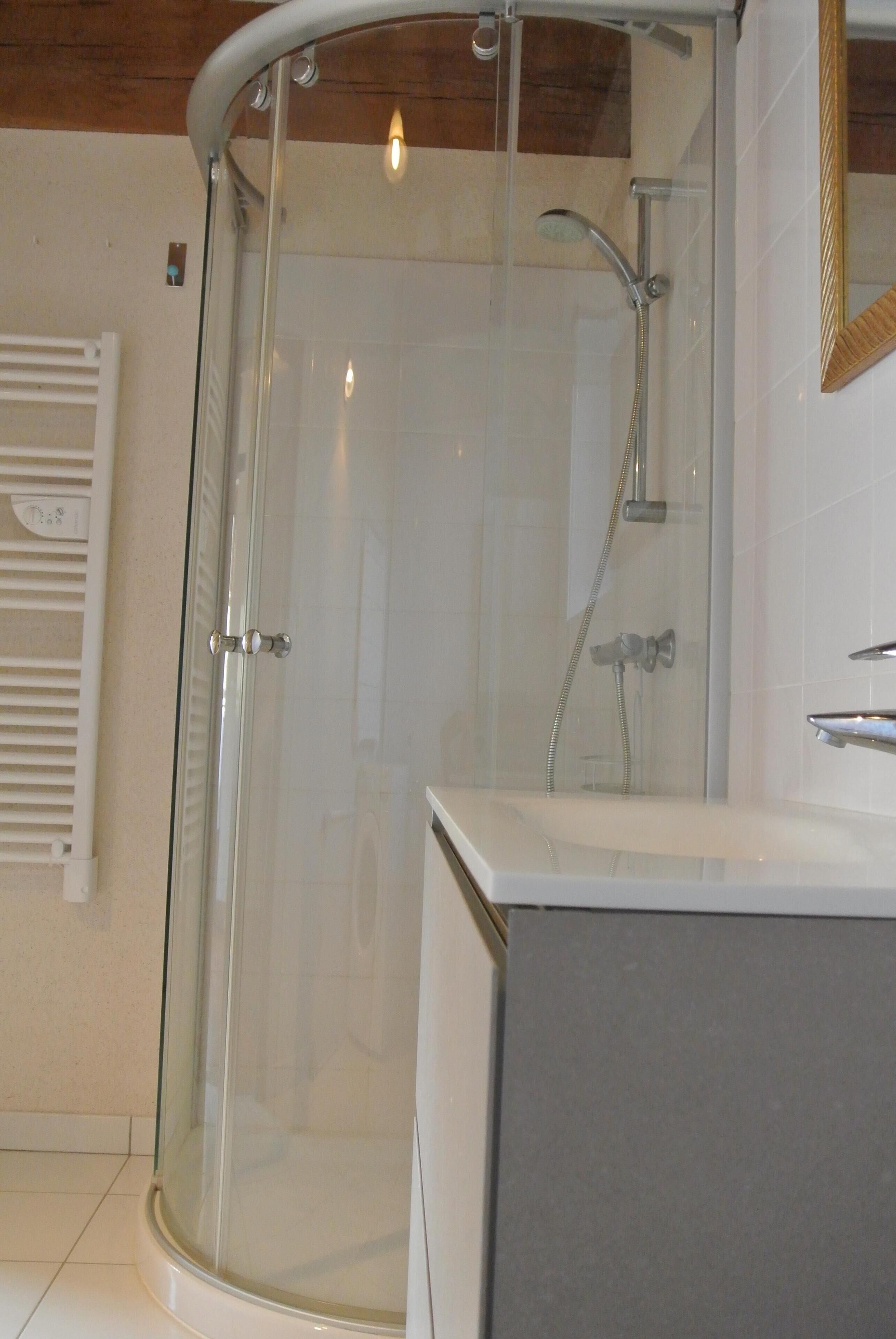 Une douche fermée par des parois de verre, dotée d’un pommeau de douche à main, d’un mur recouvert de carreaux blancs et d’un plafond en bois.