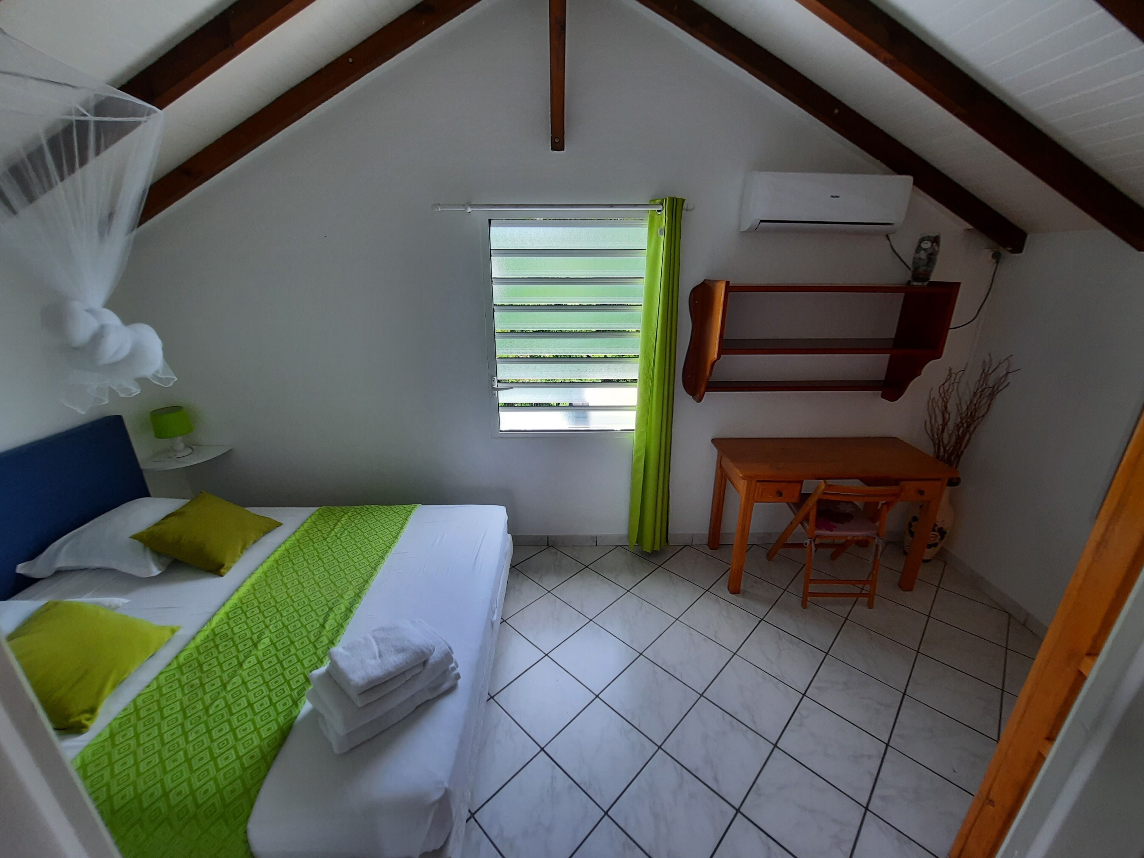 A small, well-lit room with a bed, a desk, and a window with green curtains.