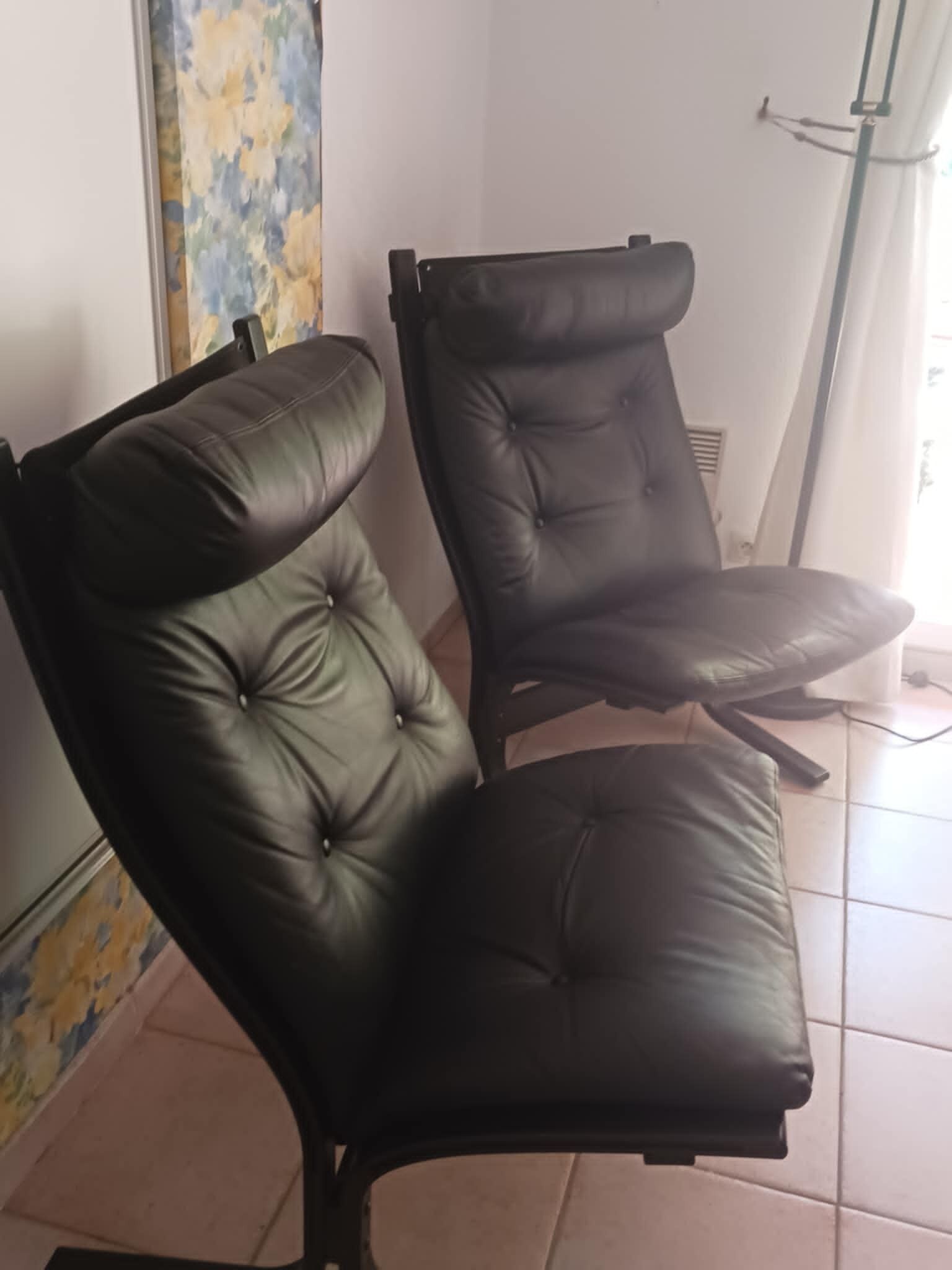 Deux fauteuils en cuir noir, dotés d’un motif capitonné, sont placés dans une pièce dont le sol est recouvert de carreaux et qui possède une fenêtre avec des rideaux.