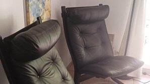 Deux fauteuils en cuir noir, dotés d’un motif capitonné, sont placés dans une pièce dont le sol est recouvert de carreaux et qui possède une fenêtre avec des rideaux.