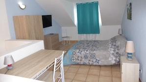 Ein kleiner, moderner Raum mit einem Bett, einem Schreibtisch und einem Sessel. Ein Fenster mit Vorhang, ein an der Wand montierter Flachbildfernseher und eine Lampe auf einem Nachttisch.