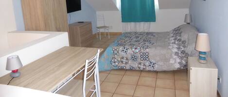 Ett litet, modernt rum med en säng, ett skrivbord och en stol. Det finns ett fönster med en gardin, en platt-tv som är monterad på väggen och en lampa på ett nattduksbord.