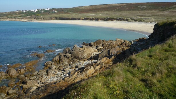 Un paisaje costero con un litoral rocoso, una playa arenosa y una colina cubierta de hierba al fondo.