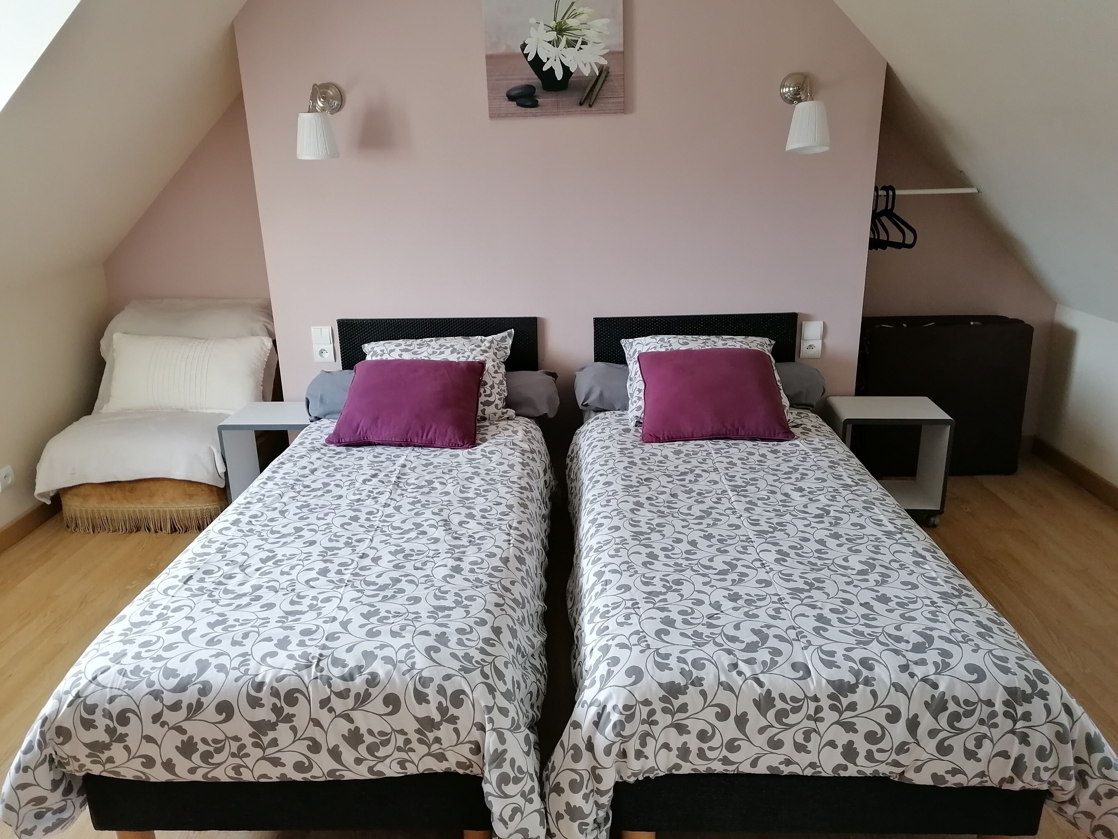 A bedroom with two beds, each with patterned bedding and pillows, a small bedside table, a nightstand, and a wall-mounted shelf with decorative items.