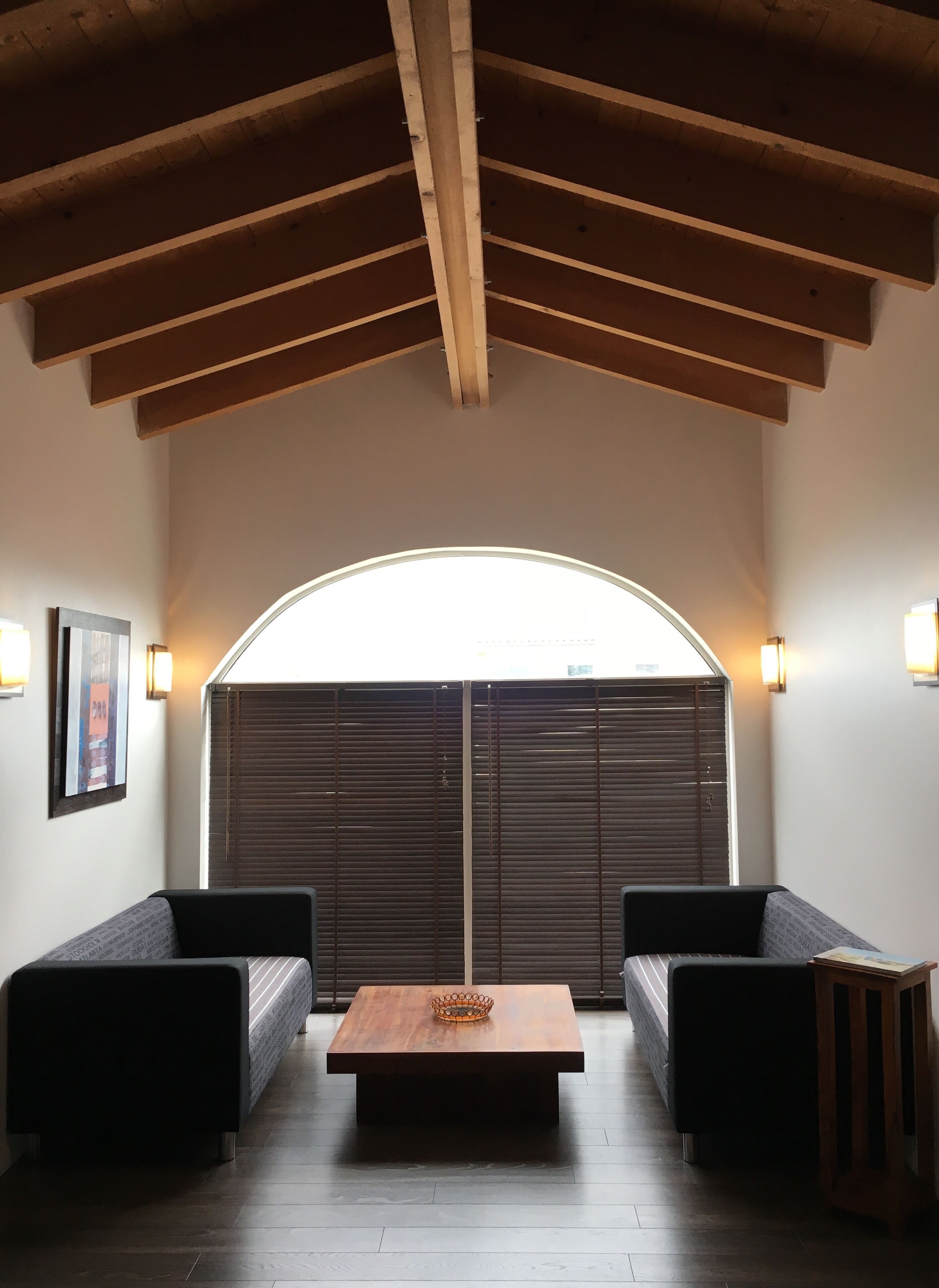 Une pièce avec un plafond en bois, une grande fenêtre avec des stores, deux canapés noirs, une table en bois et des lampes fixées au mur.