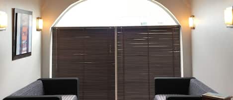 Une pièce avec un plafond en bois, une grande fenêtre avec des stores, deux canapés noirs, une table en bois et des lampes fixées au mur.