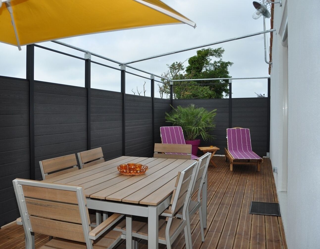 A wooden deck with a table and chairs, a yellow umbrella, and two striped lounge chairs.