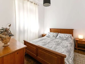 A bedroom with a wooden bed, a bedside table, a nightstand, a lamp, a window with curtains, and a vase with dried plants on a wooden dresser.