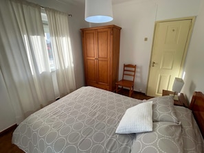 A bedroom with a bed, a wooden wardrobe, a chair, a door, and a window with curtains.