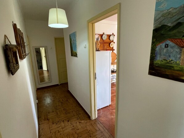 Un pasillo con suelo de madera, una puerta blanca y un refrigerador blanco. Hay un colgante de luz y un cuadro en la pared.