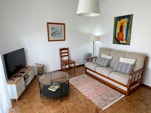 Una sala de estar con un sofá, un televisor, una mesa de centro de vidrio, una silla, una lámpara y un cuadro en la pared.