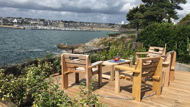 Eine Holzterrasse mit Tisch und Stühlen, die auf einen Yachthafen mit Booten und eine Küstenstadt im Hintergrund blickt.