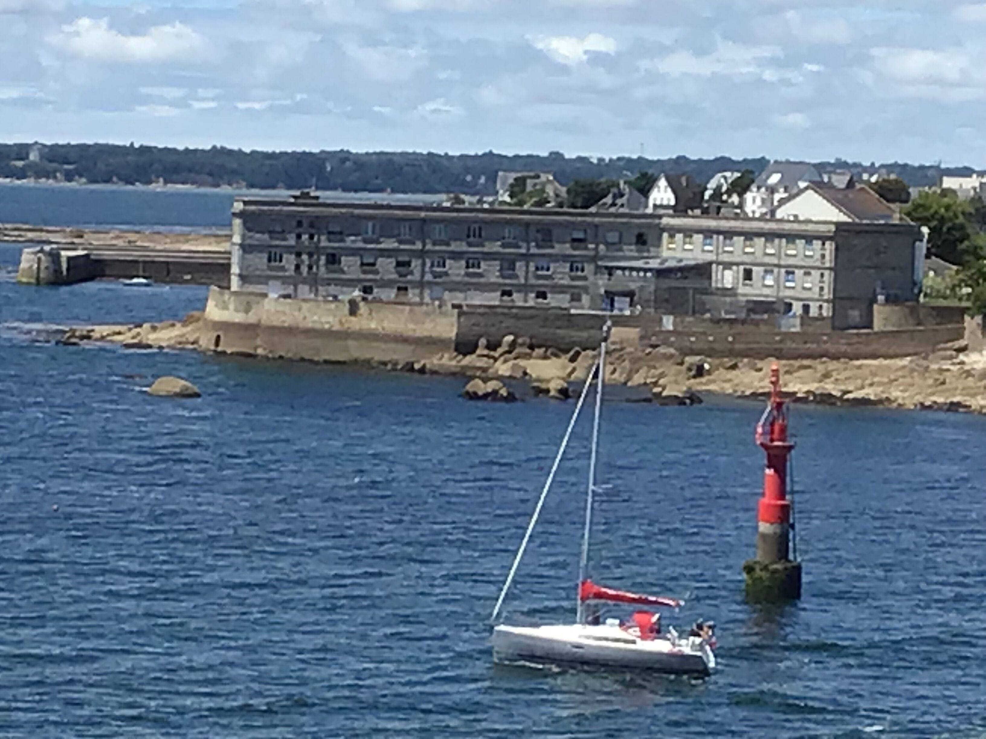 A sailboat on calm waters near a lighthouse and a building with multiple windows.