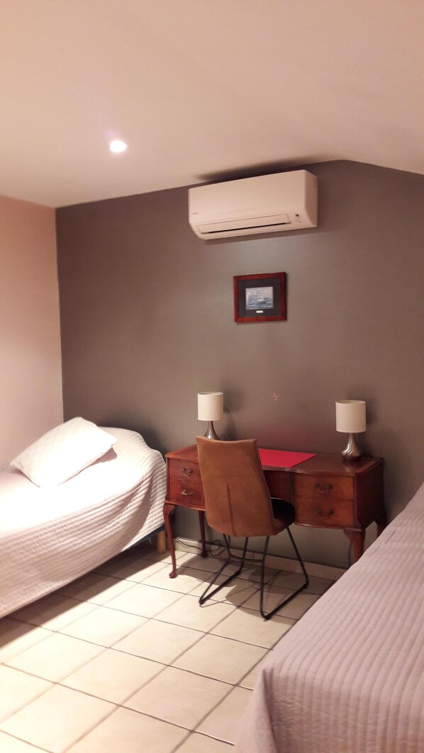 Un dormitorio con cama, escritorio con dos lámparas, silla, mesita pequeña y aire acondicionado.