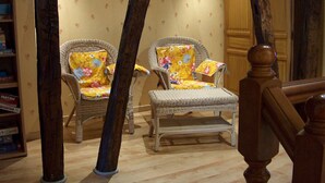 A room with two wicker chairs, one with a floral cushion and another with a plain cushion. There is a wooden bookshelf with books and a wooden door in the background.