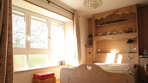 Ein Schlafzimmer mit einem hölzernen Bett, einer Bank, einem Fenster mit Vorhängen und einem Regal mit Dekoration.