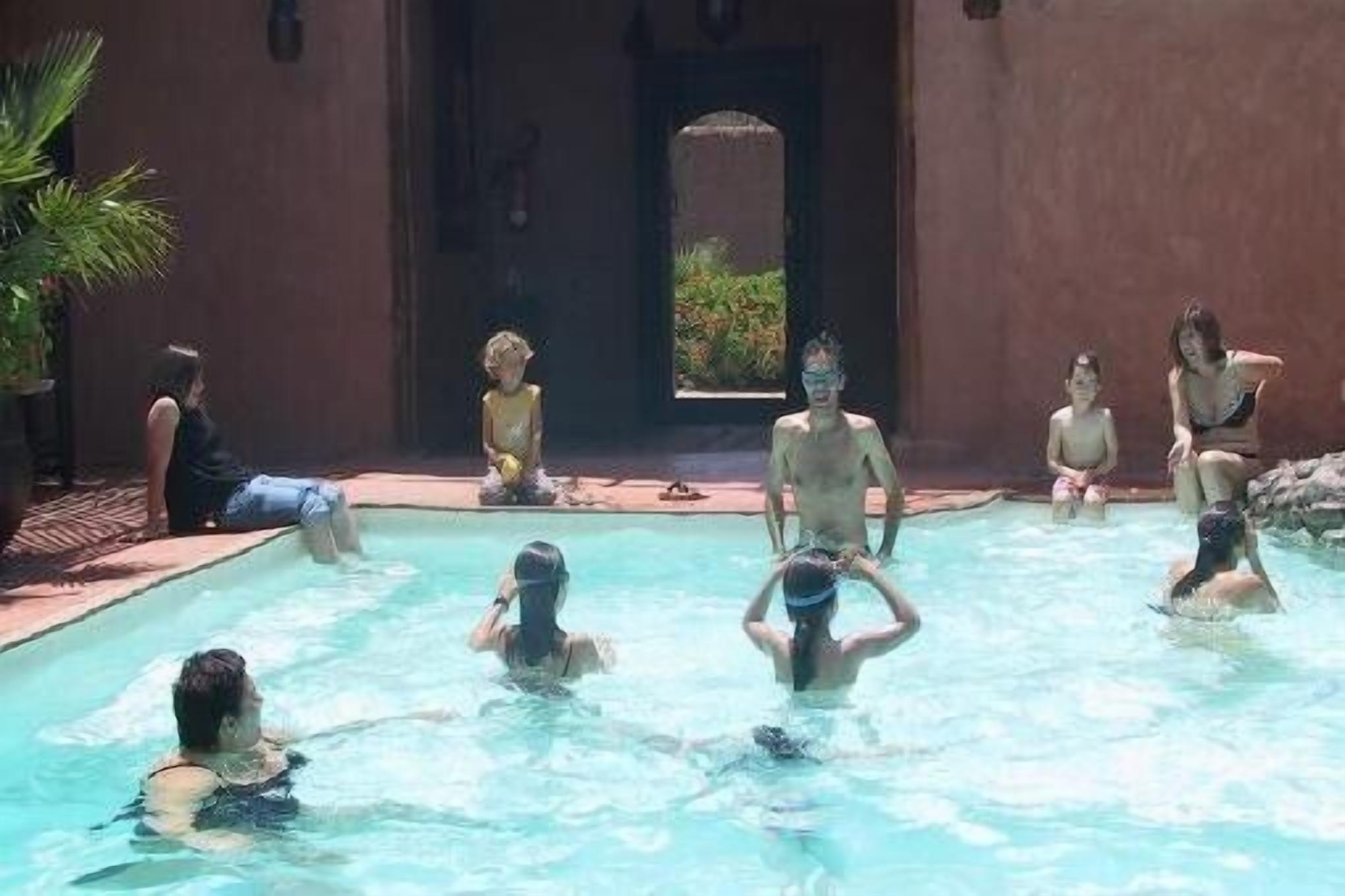 Une zone autour d’une piscine, avec des personnes dans l’eau et d’autres assises au bord.