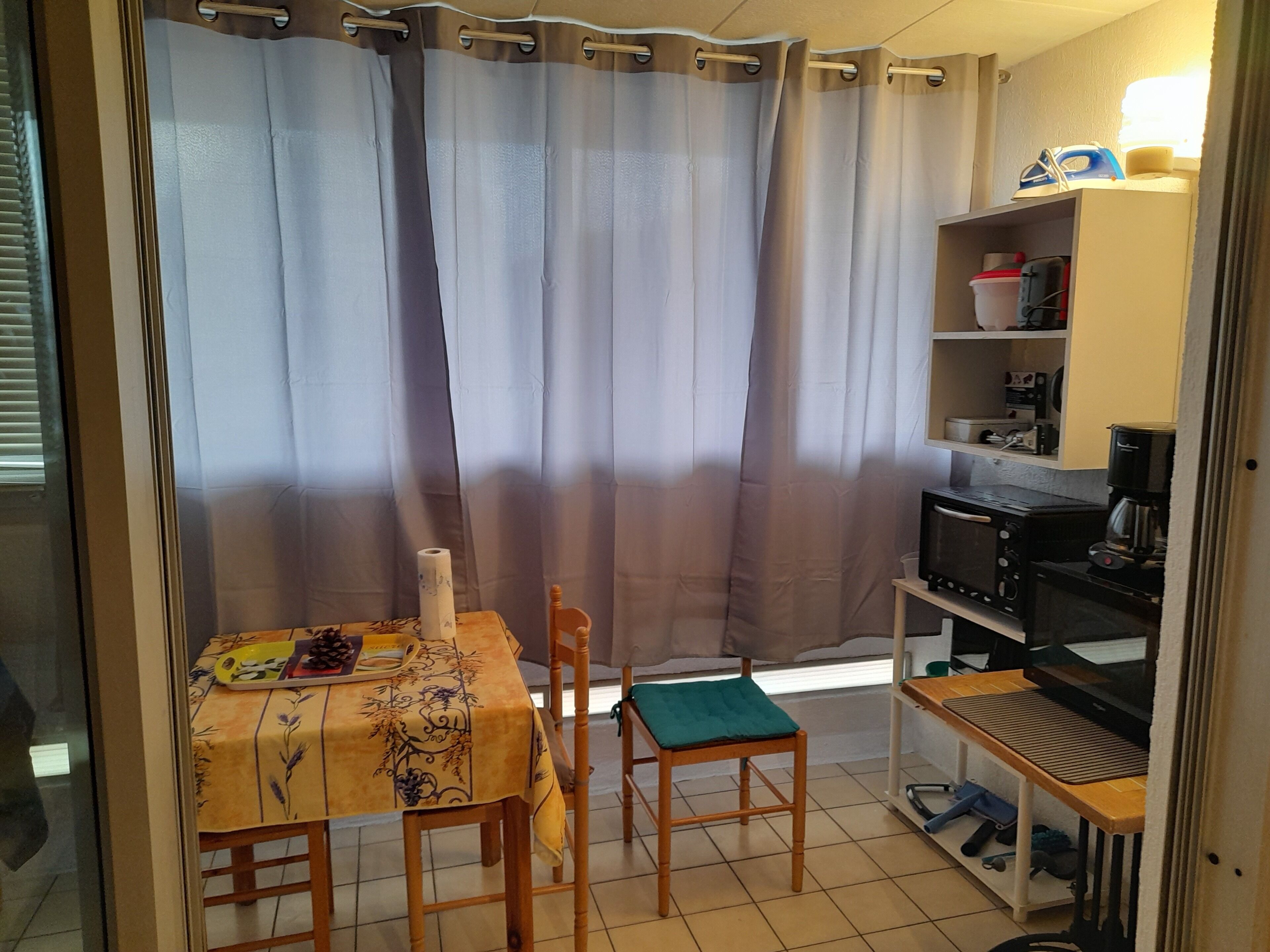 A small kitchenette with a coffee maker, toaster, and shelves with kitchenware. There is a table with a floral tablecloth and two chairs.