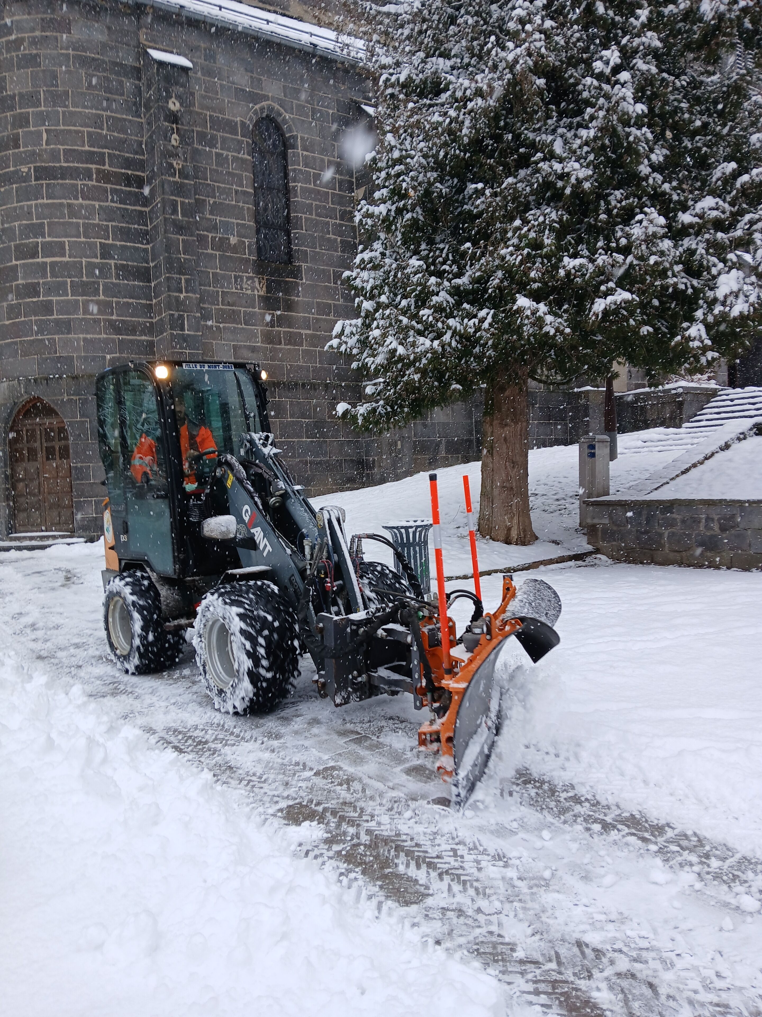 Un chasse-neige devant un bâtiment en briques, avec un arbre.