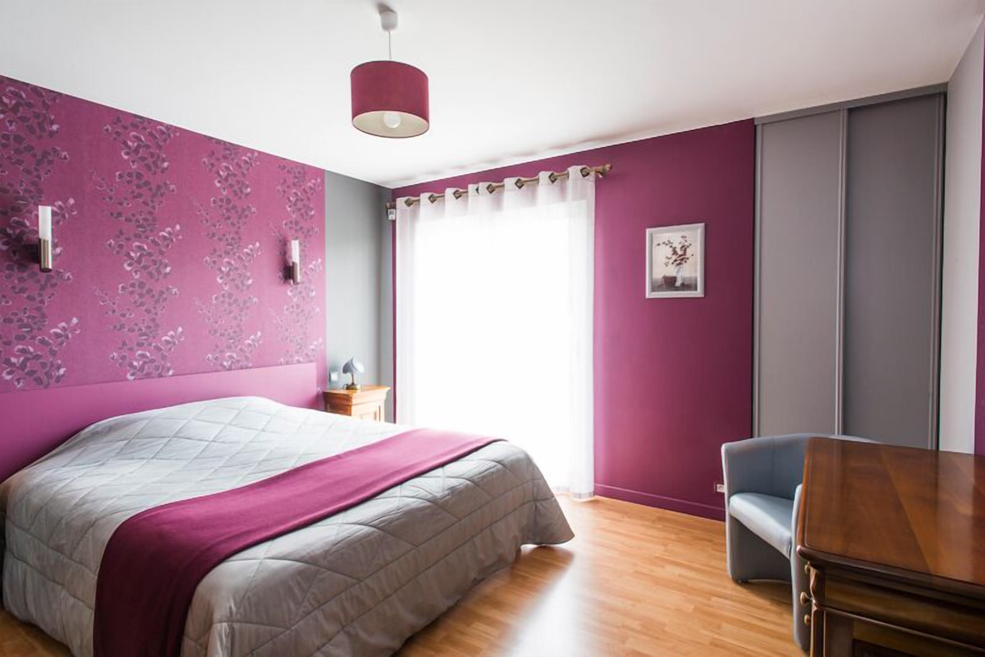 Un dormitorio con una cama, una mesa de madera, una silla, un ventanal con cortinas y una pared con estampado floral.