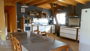 A dining area with a table set for six, wooden chairs, a kitchen in the background with appliances like a microwave, stove, and refrigerator, and a wooden ceiling structure.