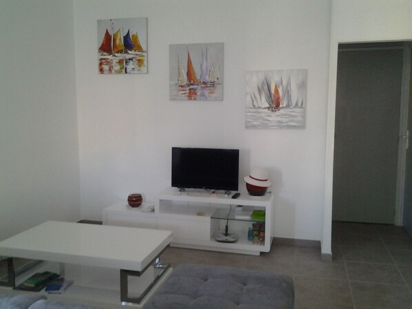 A living room with a flat-screen TV on a white stand, a white coffee table, a grey sofa, and three paintings on the wall. - HIGH T3 APARTMENT VILLA of 60 m2 (Sanary-Sur-Mer)
