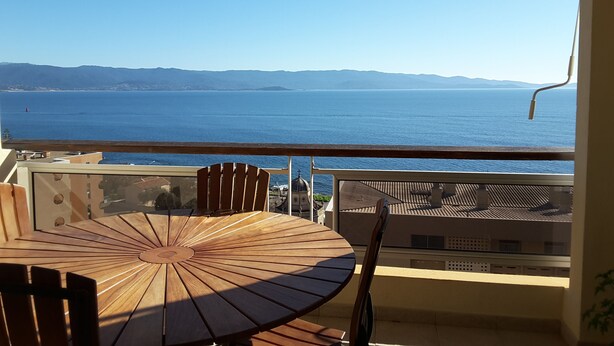 A wooden table and chairs on a balcony overlooking a large body of water with distant mountains.