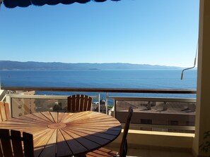 Una mesa y sillas de madera en un balcón con vista a una extensa masa de agua y montañas a lo lejos.