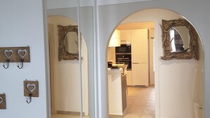 A hallway with white walls, a mirrored wardrobe, and a kitchen area visible through an archway.