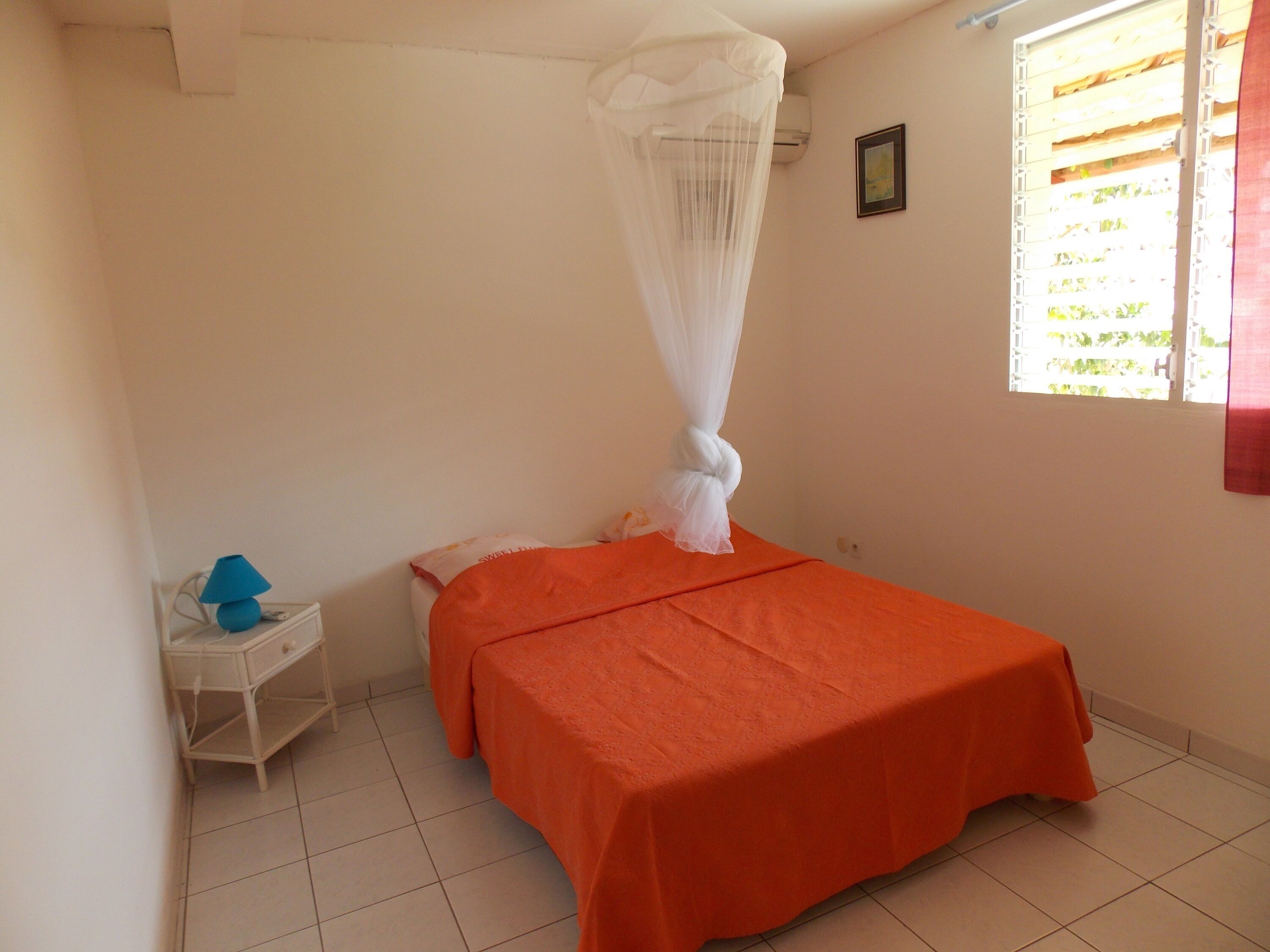A bedroom with a bed covered in an orange bedspread, a white nightstand with a blue lamp, and a window with a white curtain.