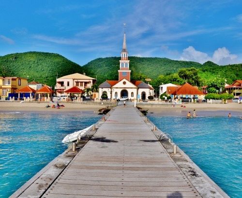 Un muelle de madera que da a una playa, con una iglesia al fondo y montañas.