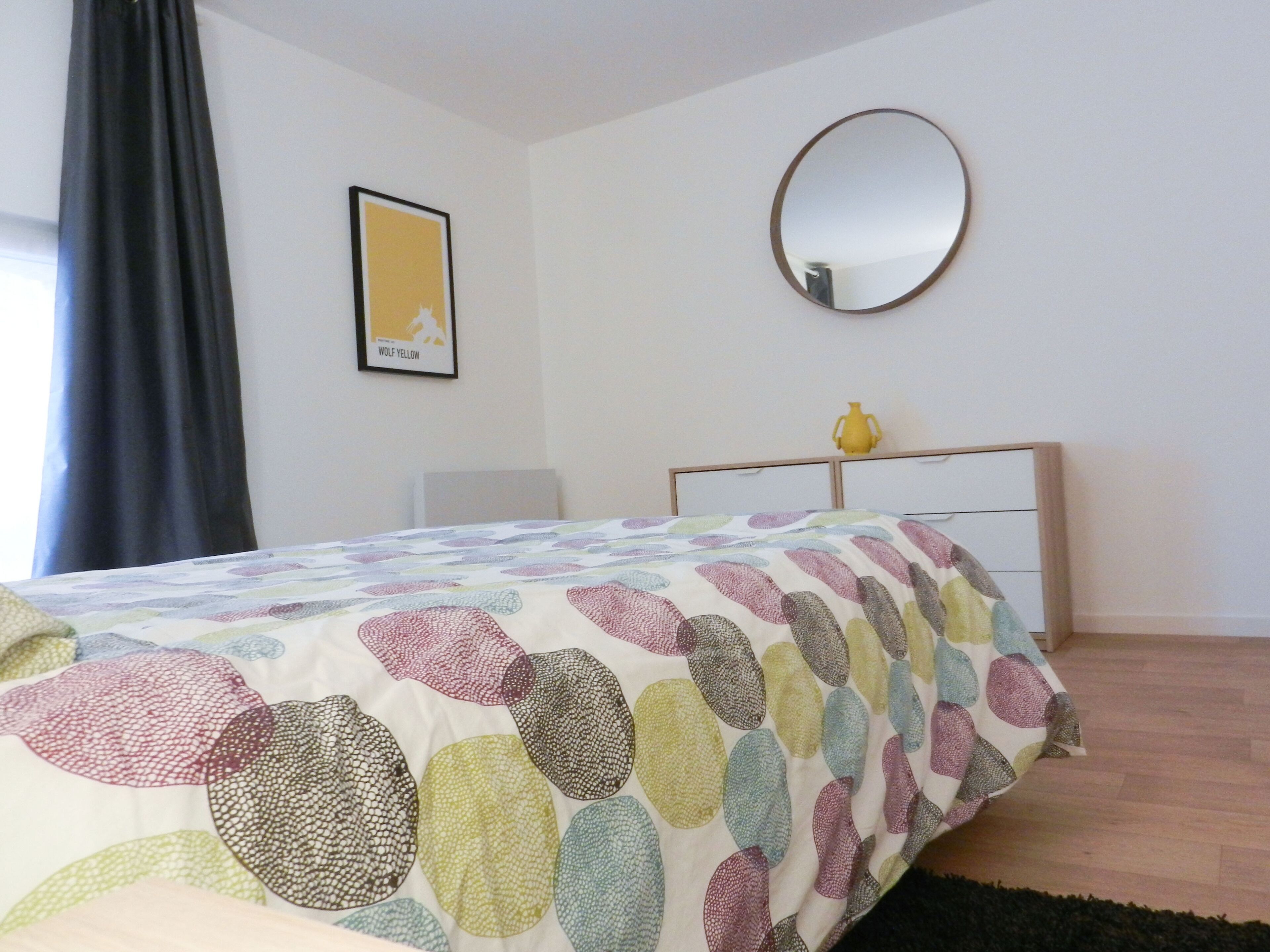 A bedroom with a bed, a bedside table, a mirror, a framed picture, and a window with curtains.