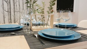 Una mesa de comedor dispuesta con platos azules, vasos transparentes y cubiertos, rodeada de pequeños jarrones de vidrio con follaje.