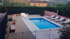Una piscina rectangular con un borde de baldosas, rodeada por un muro de ladrillo bajo. Hay sillas de descanso y una pequeña mesa con una planta en maceta. Se observa un edificio con techo de tejas al fondo.
