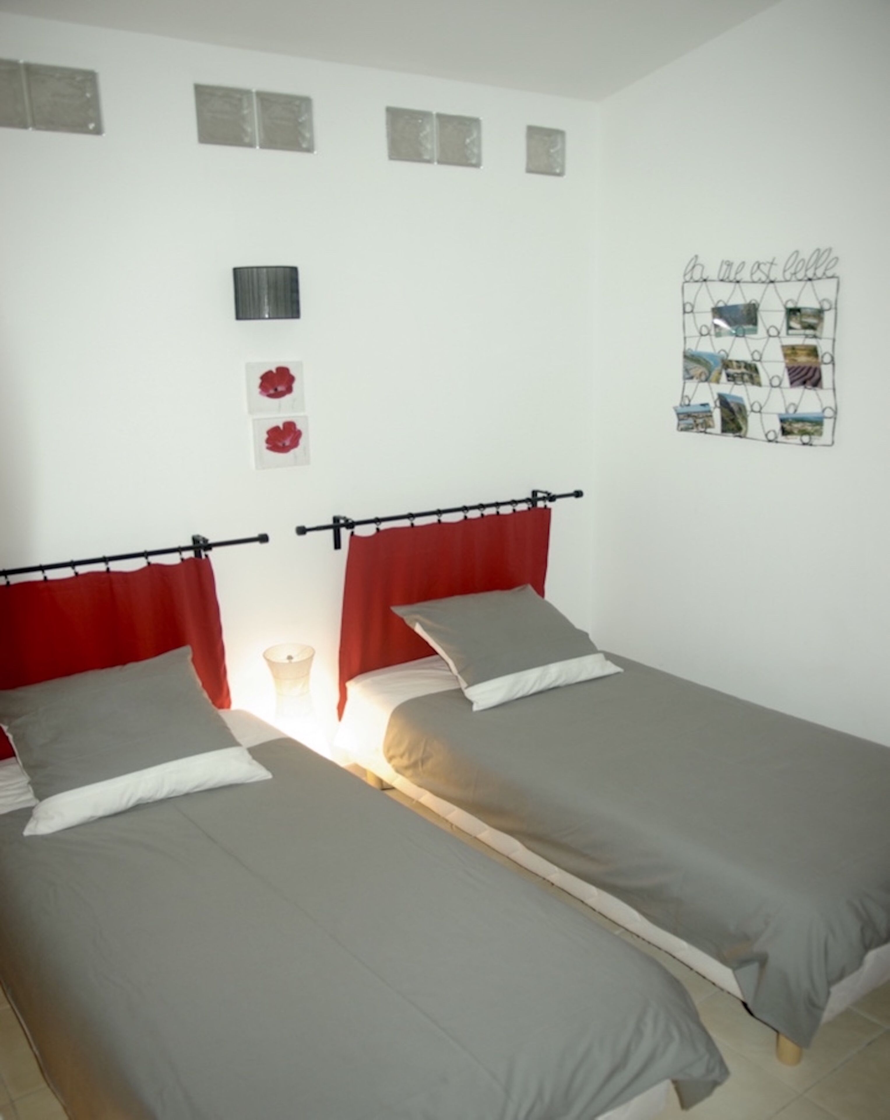 A room with two single beds, each with a red headboard and grey bedding. There is a wall-mounted shelf with decorative items and a framed collage.