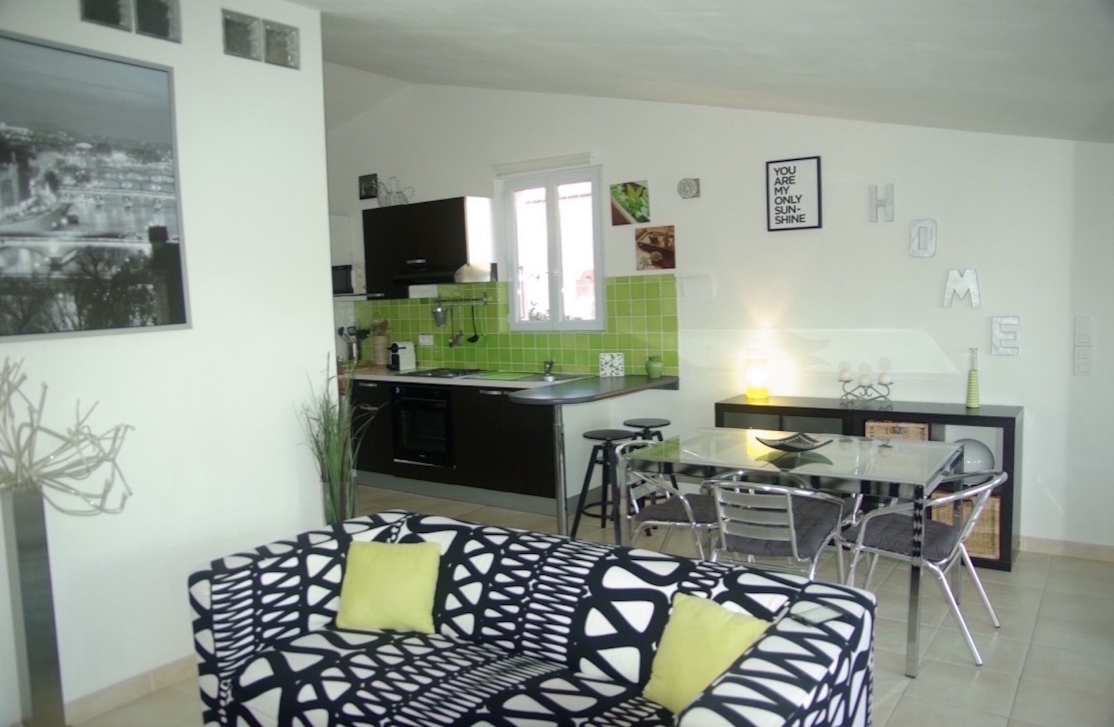 A modern kitchen with a green backsplash, a dining area with a glass table and metal chairs, and a black and white patterned sofa with green cushions.